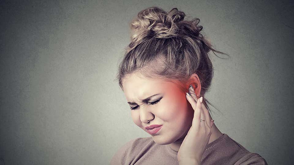 Uma pessoa, com o rosto desfocado, usa discretamente um aparelho auditivo atrás da orelha, enquanto toca a lateral da cabeça. O fundo é neutro e uniforme.