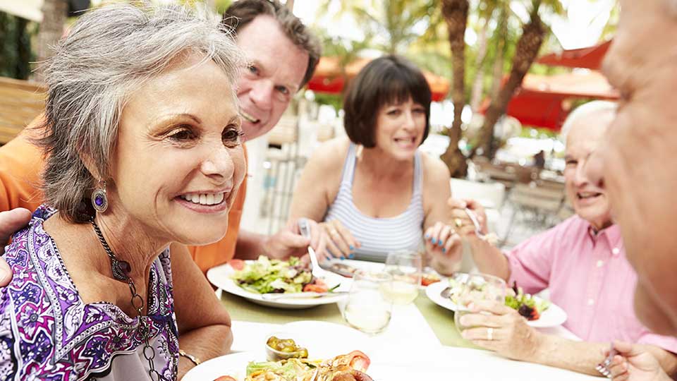 Idosos reunidos ao redor de uma mesa ao ar livre, compartilhando uma refeição em um ambiente tropical com guarda-sóis vermelhos, pratos variados e taças de bebida, sugerindo um momento social descontraído.