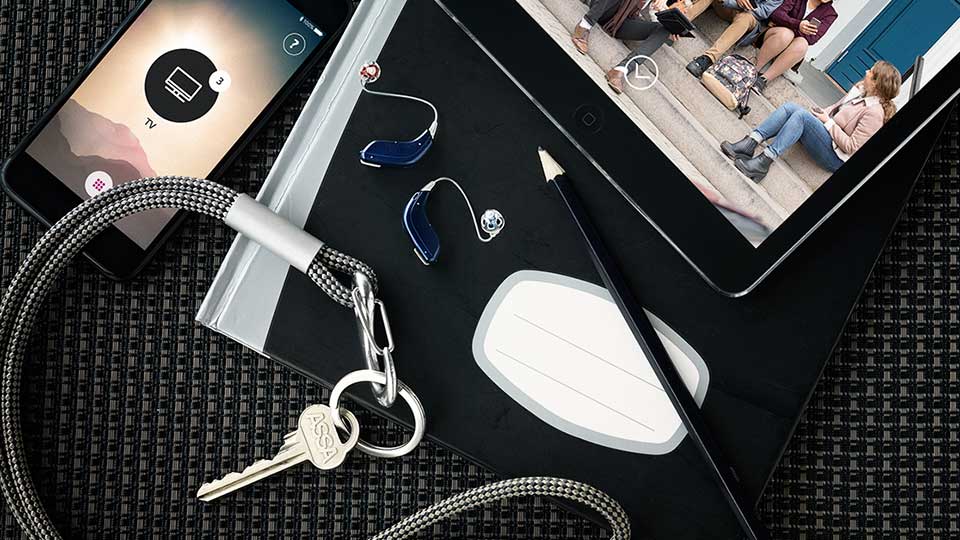 Dois aparelhos auditivos azul e prata da Oticon posam sobre um caderno preto ao lado de um tablet mostrando pessoas conversando, um smartphone exibindo 'TV' e um chaveiro prateado.