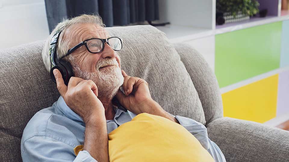 Idoso relaxando em um sofá com uns auscultadores, com uma almofada amarela apoiada no colo.