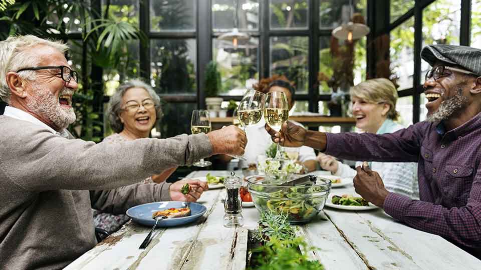 Pessoas idosas brindando em uma refeição comunitária com pratos saudáveis em um ambiente cheio de plantas.