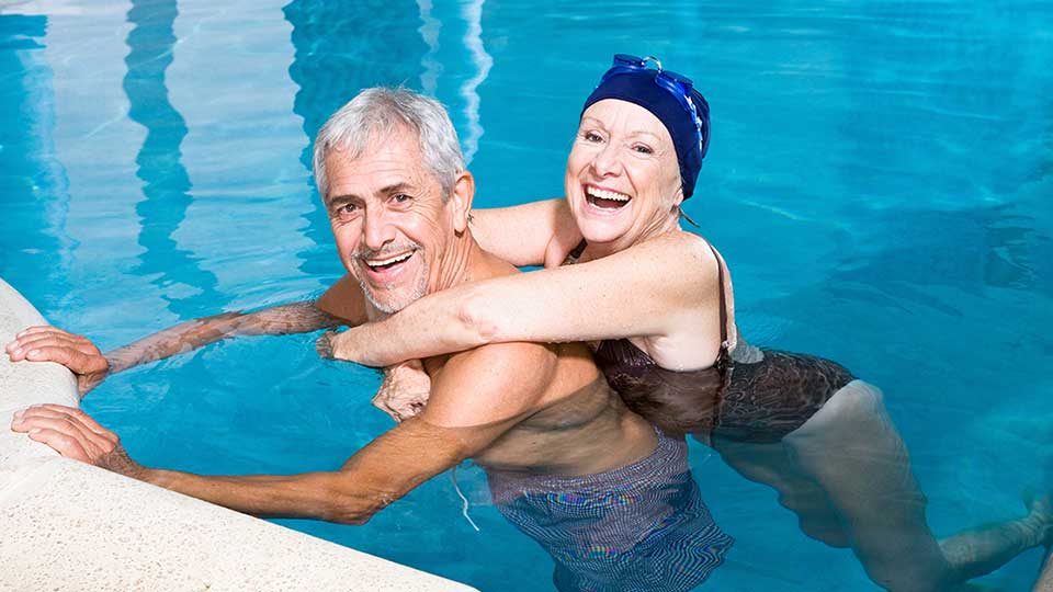 Um casal idoso está em uma piscina; a mulher usa touca azul, abraçando o homem que se apoia na borda. O ambiente sugere lazer e relaxamento.