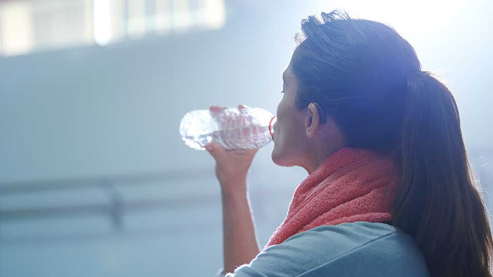 Mulher a beber água após o exercício, com toalha ao redor do pescoço.