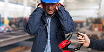 Um trabalhador usando capacete azul cobre os ouvidos com as mãos em um ambiente industrial desfocado, enquanto uma outra pessoa oferece protetores auriculares vermelhos para proteção auditiva no local.