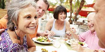 Um grupo de pessoas idosas está reunido em um restaurante ao ar livre, compartilhando uma refeição. As expressões parecem descontraídas e amistosas, em um ambiente ensolarado com guarda-sóis vermelhos ao fundo.