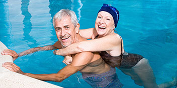 Duas pessoas idosas em trajes de banho estão em uma piscina, com uma delas abraçando a outra. O ambiente de lazer é cercado por água azul clara, típico de piscina recreativa.
