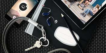 Dois aparelhos auditivos azuis Oticon estão pousados sobre um caderno preto ao lado de um lápis, chaveiro com chave, smartphone exibindo um aplicativo e tablet mostrando uma cena urbana.