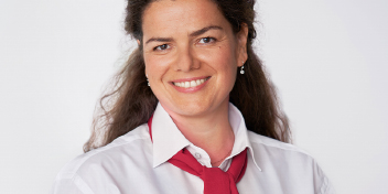 Mulher com rosto desfocado veste camisa branca e gravata vermelha solta. O fundo é branco, sugerindo um ambiente profissional ou promocional, possivelmente relacionado à saúde auditiva ou atendimento em clínica.