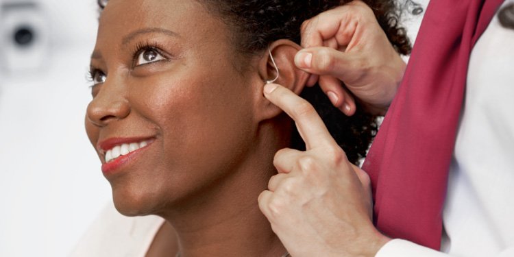 Um audiologista ajusta um aparelho auditivo discreto atrás da orelha de um paciente num centro auditivo especializado, com fundo branco desfocado sugerindo ambiente profissional. Texto ausente.