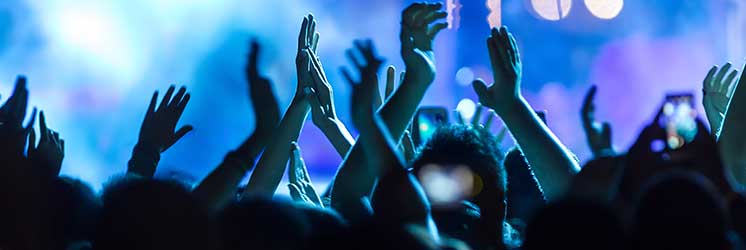 Mãos erguidas aplaudem em um show ou festival, com luzes azuis e púrpuras iluminando o ambiente noturno repleto de espectadores, alguns segurando celulares para registrar o momento.