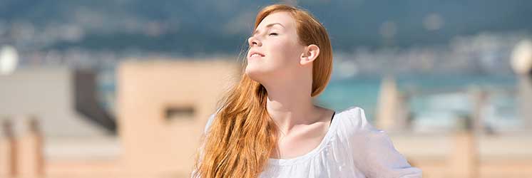 Uma pessoa com cabelo ruivo longo está ao ar livre usando uma blusa branca. Ao fundo, prédios desfocados e um cenário costeiro com montanhas ao longe. Sem texto visível.