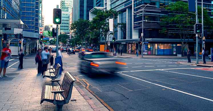 Bancos de praça alinhados enquanto pedestres caminham e carros atravessam um cruzamento urbano movimentado com semáforos verdes. Cercado por modernos prédios envidraçados e árvores, criando um ambiente de cidade contemporânea.