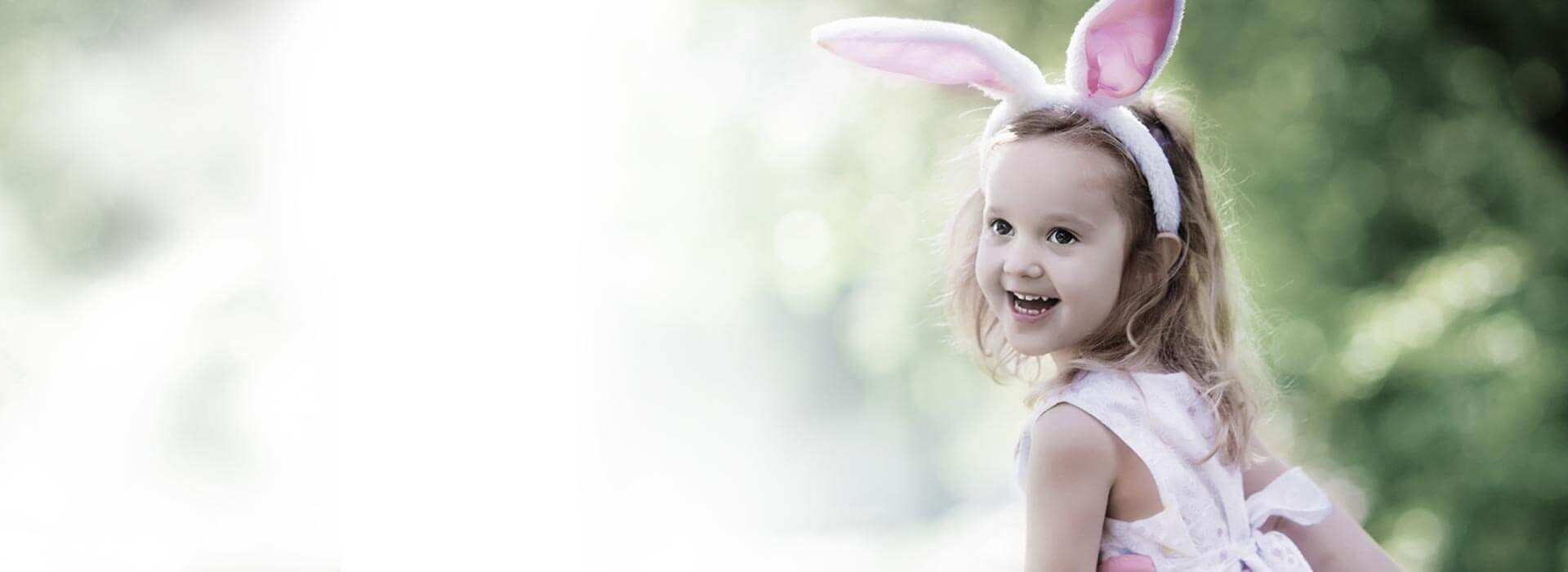 Uma criança com faixa de orelhas de coelho está sentada ao ar livre, cercada por um fundo desfocado de folhagem verde, iluminado por luz natural suave.