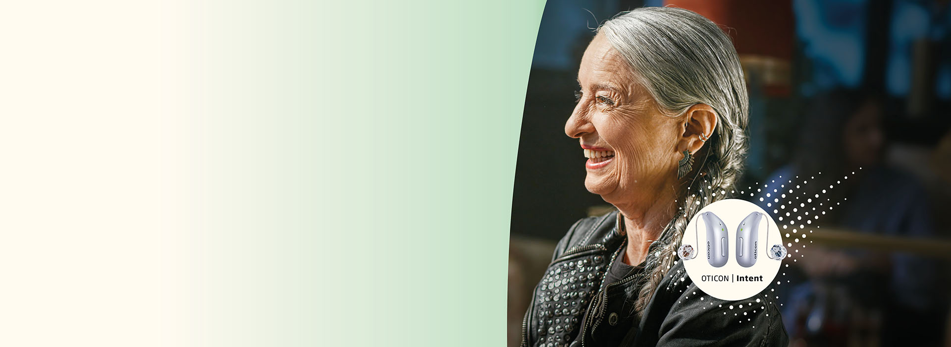 Mulher de cabelos grisalhos com jaqueta de couro sentada em ambiente interno; destaque para aparelhos auditivos ’OTICON | Intent’ em um círculo no canto inferior direito da imagem.