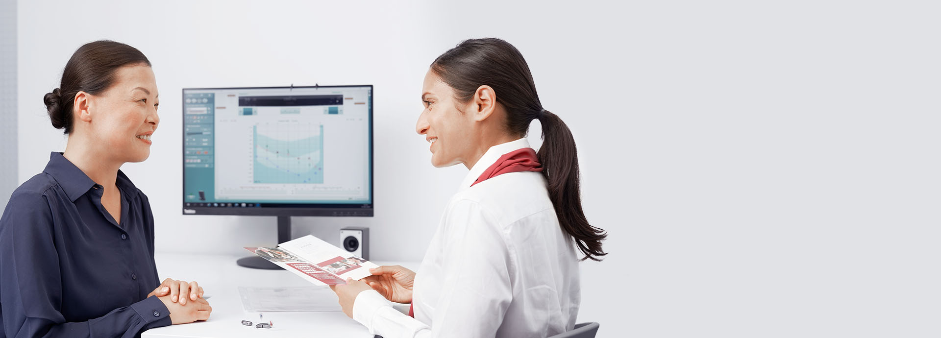 Cliente e audiologista interagem em uma clínica auditiva. A audiologista, segurando um folheto informativo, explica informações enquanto um audiograma aparece na tela do computador ao fundo. Aparelhos auditivos estão sobre a mesa.