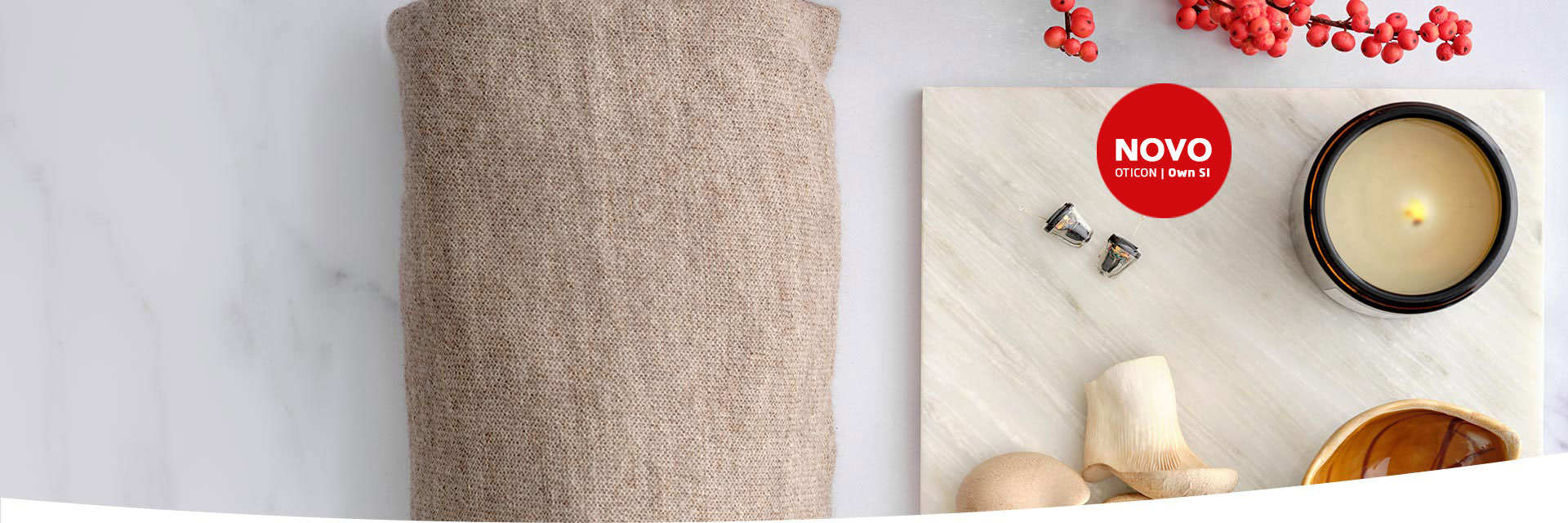 Aparelhos auditivos intra-auriculares Oticon | Own SI estão sobre uma superfície clara junto a uma vela acesa, decoração natural e tecido bege; texto destacado: 'NOVO Oticon | Own SI'.