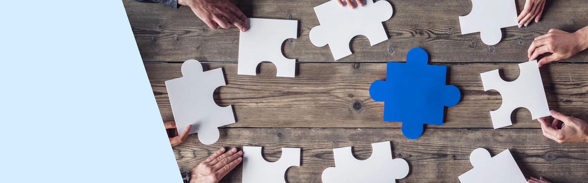 Peças de quebra-cabeça brancas e uma azul são dispostas sobre uma mesa de madeira, enquanto mãos colaboram para posicioná-las no lugar correto, sugerindo trabalho em equipe ou resolução conjunta.