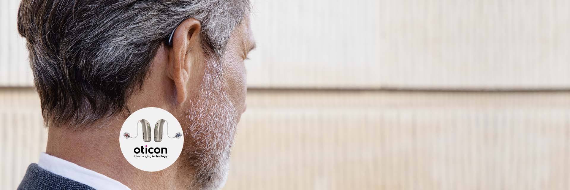 Um aparelho auditivo da marca Oticon está encaixado discretamente atrás da orelha de um homem grisalho. Fundo desfocado em tons claros. Texto: ’Oticon, life-changing technology’ com imagem de três aparelhos auditivos.