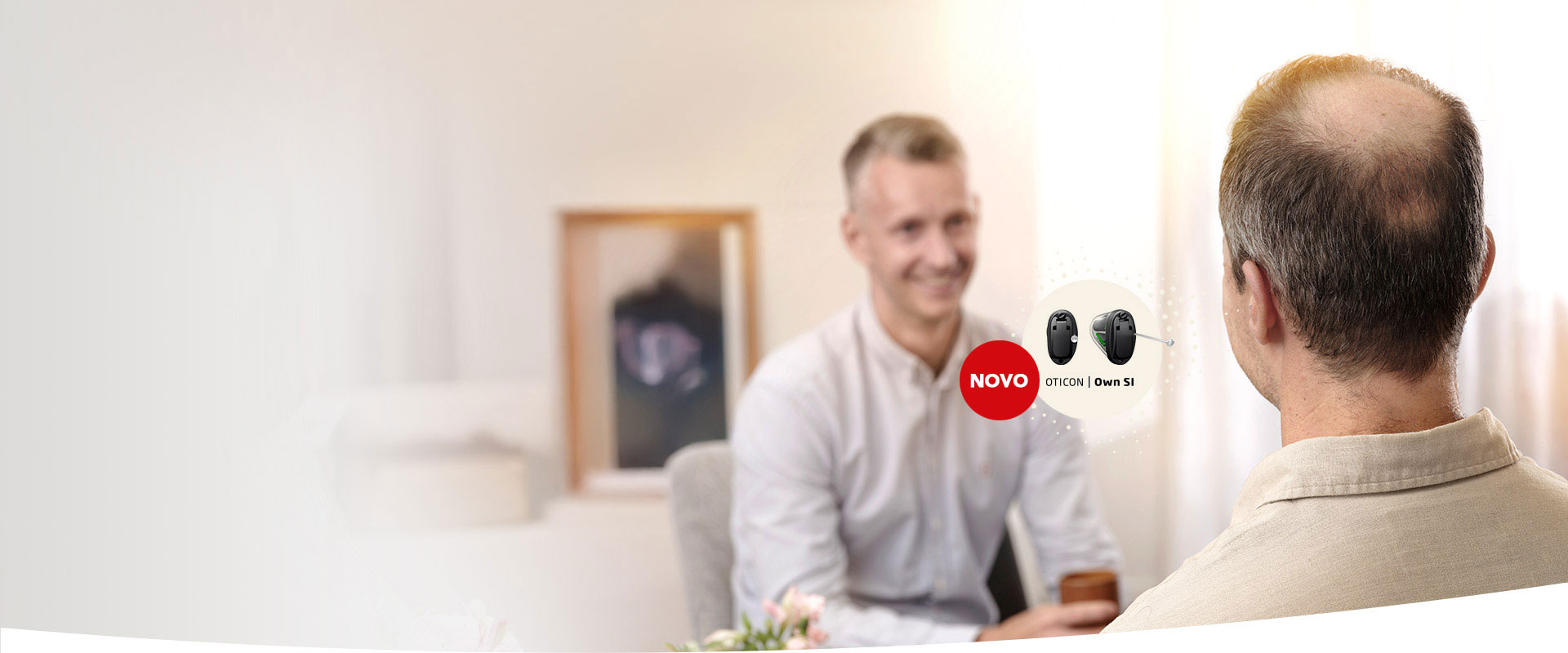 Homem calvo de costas conversando com um profissional num centro auditivo iluminado e aconchegante. À direita, destaque para aparelhos auditivos ’Novo Oticon Own S1’ com design discreto.