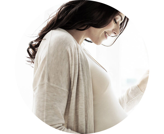 Mulher grávida, vestindo cardigan e camiseta clara, posicionada de perfil enquanto segura suavemente a barriga. O fundo é iluminado por luz natural, sugerindo um ambiente interno arejado.