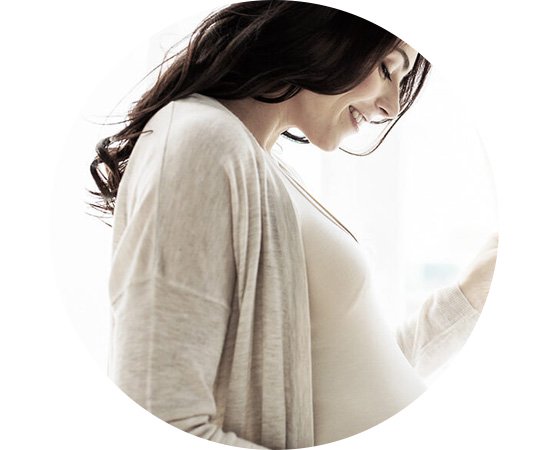 Mulher grávida, vestindo cardigan e camiseta clara, posicionada de perfil enquanto segura suavemente a barriga. O fundo é iluminado por luz natural, sugerindo um ambiente interno arejado.