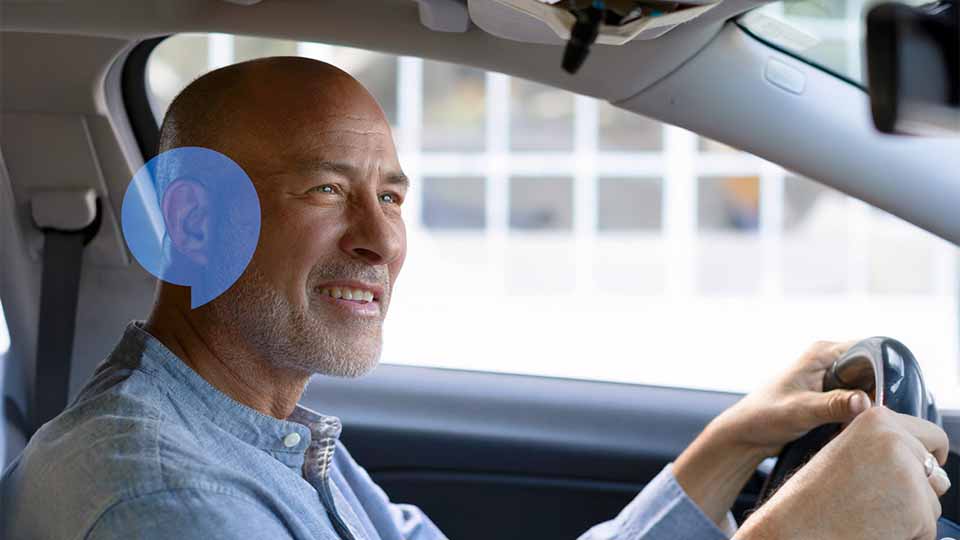 Un homme conduit une voiture, portant un appareil auditif discret. Le contexte montre un environnement urbain flou à travers la vitre. Aucune marque spécifique n’est identifiable.