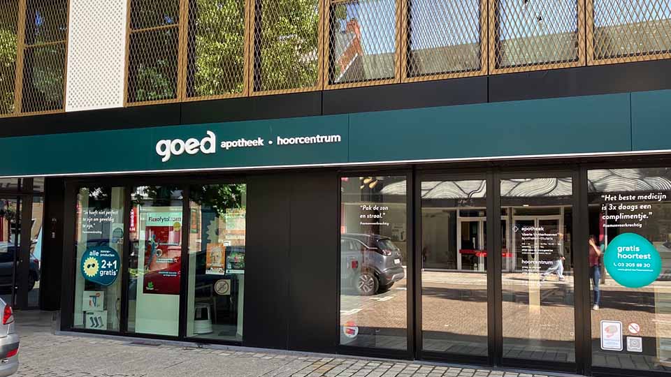 Une façade de bâtiment moderne affiche 'goed apotheek - hoorcentrum' sur une enseigne verte. Une vitrine mentionne 'gratis hoortest' (test auditif gratuit) accompagné d'offres et de décoration intérieure pharmaceutique.