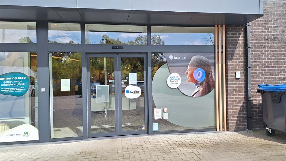 Façade d'une clinique Audika avec portes vitrées automatiques. Affiche promotionnelle montrant une femme portant un appareil auditif. Texte visible : ''Audika'', ''gratis en vrijblijvend hoortoestel'', et panneaux explicatifs en néerlandais.