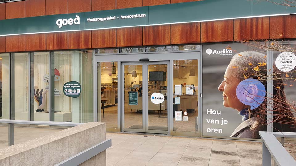 La façade d'un centre auditif combiné à une boutique 'goed thuiszorgwinkel - hoorcentrum.' Action : portes vitrées ouvertes sur l'intérieur clair. Texte : 'Audika Hoorcentrum,' 'Hou van je oren,' et 'gratis en vrijblijvende hoortest binnen 30 min.' Contexte : environnement urbain.