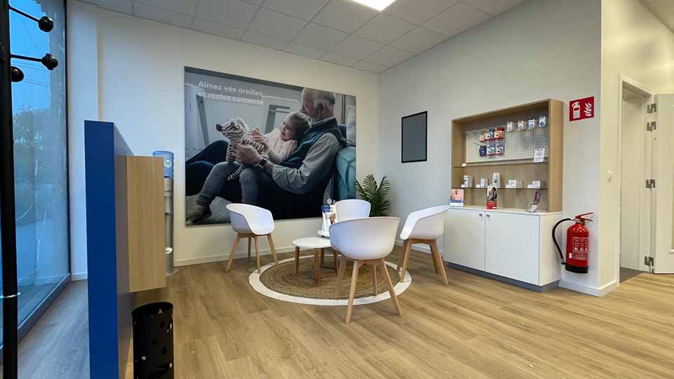 Une salle d'attente lumineuse d'une clinique auditive Audika, équipée de chaises blanches autour d'une table, une affiche murale 'Aimez vos oreilles et restez connecté', étagères de produits auditifs, extincteur rouge à droite.