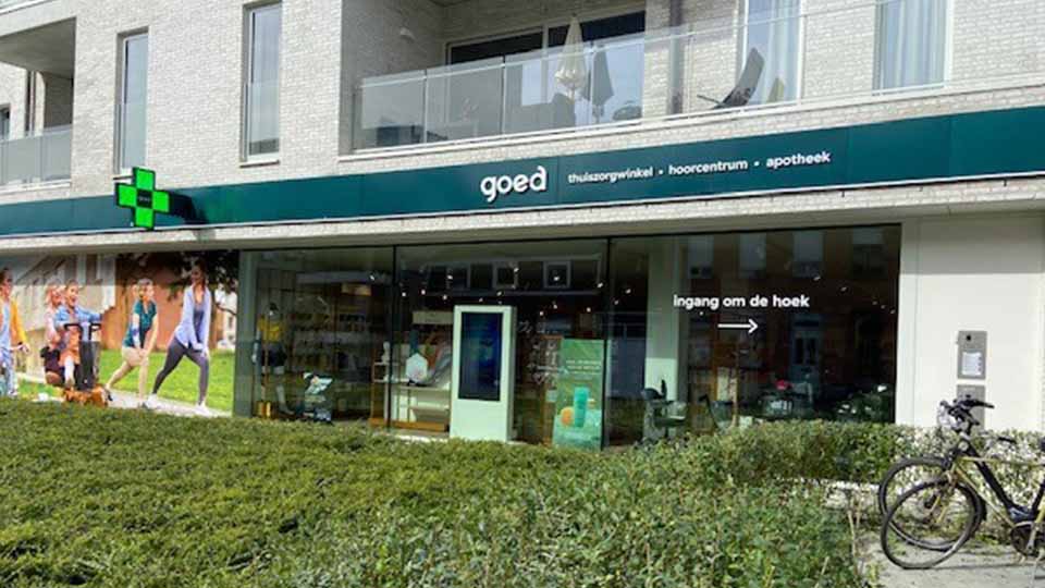 Une façade de magasin moderne avec une enseigne verte 'goed' mentionnant 'thuiszorgwinkel • hoorcentrum • apotheek'. À droite, une flèche indique 'ingang om de hoek'. Un vélo est stationné devant.