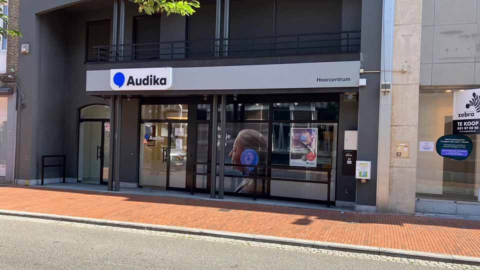 La façade d'un centre auditif Audika avec enseigne blanche et bleue, marqué 'Hoorcentrum'. Fenêtres vitrées affichent des publicités sur les solutions auditives. Situé sur un trottoir urbain.
