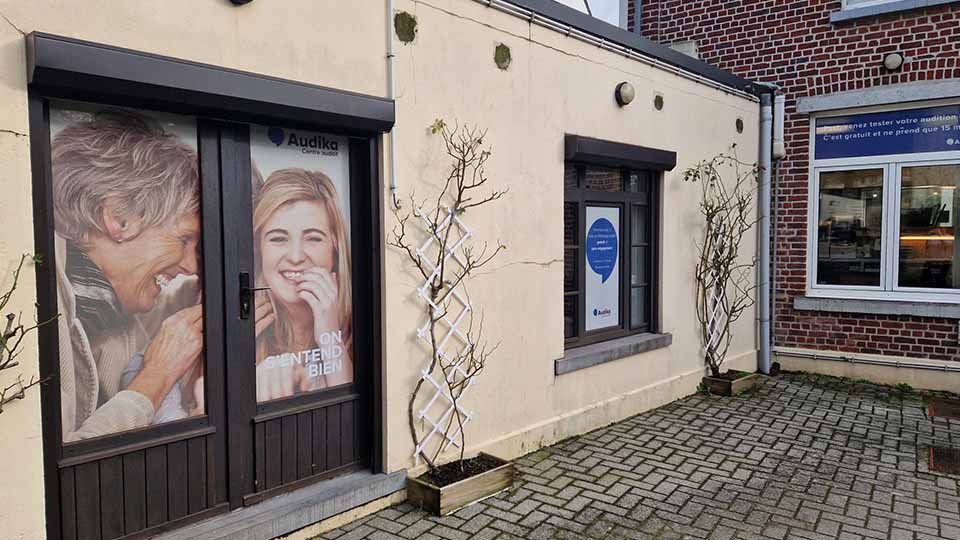 Les portes vitrées d'une clinique Audika affichent un visuel promotionnel partiellement flouté et le slogan 'On entend bien'. Un panneau encourage à tester l’audition gratuitement. Environnement pavé et murs extérieurs clairs.