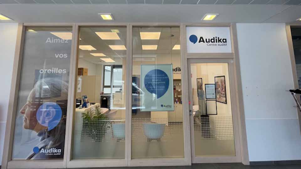 Façade d’un centre auditif Audika avec une entrée vitrée affichant le logo ’Audika Centre auditif’. Texte visible : ’Aimez vos oreilles’. Intérieur moderne avec mobilier et éclairage lumineux.