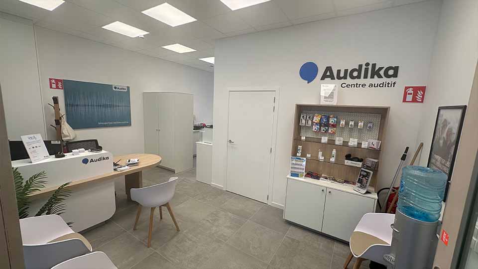 Un centre auditif Audika lumineux avec comptoir d’accueil, chaises blanches, étagère présentant des accessoires auditifs, affiche Oticon au mur, distributeur d’eau et signalisation de sécurité.