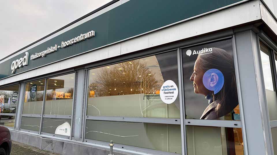 Une façade de bâtiment avec enseignes ’Goed thuiszorgwinkel - hoorcentrum’ et ’Audika hoorcentrum’. Une vitrine affiche une oreille équipée d’une aide auditive. Texte : ’gratis en vrijblijvende hoortest (0800 35 89)’.