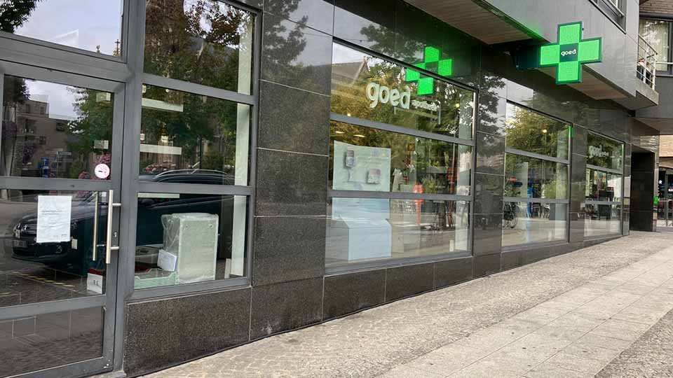 La façade d’une pharmacie avec des vitres modernes, un logo croix verte allumé indiquant ’goed’, et des reflets d’arbres environnants. Texte affiché : ’goed apotheek’.
