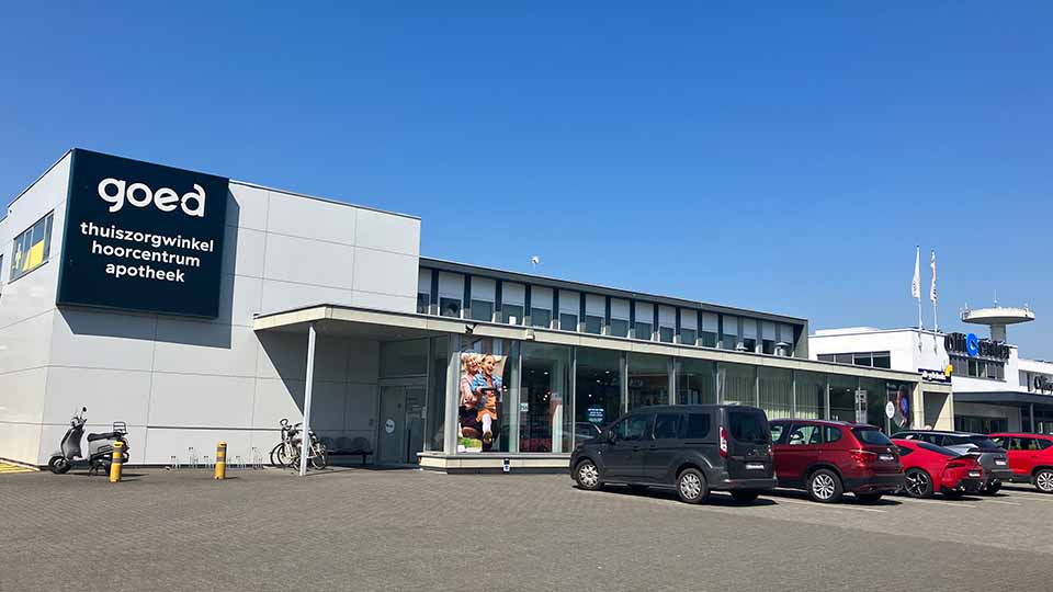 Un bâtiment moderne avec le logo ’goed’ affichant les services ’thuiszorgwinkel, hoorcentrum, apotheek.’ Des voitures sont garées devant, et le ciel est clair.