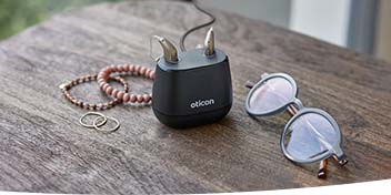 Appareil auditif Oticon sur un chargeur avec des lunettes et des bijoux posés sur une table en bois.