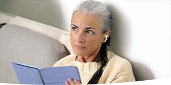 Femme âgée assise lisant un livre avec des écouteurs blancs.