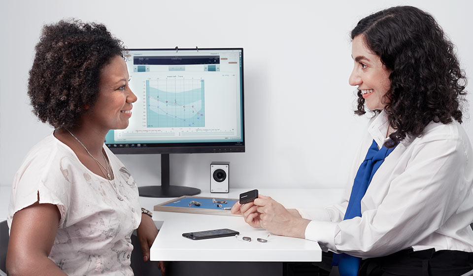 Une audiologue expliquant des accessoires pour appareils auditifs à une patiente avec un audiogramme affiché sur l'écran en arrière-plan.