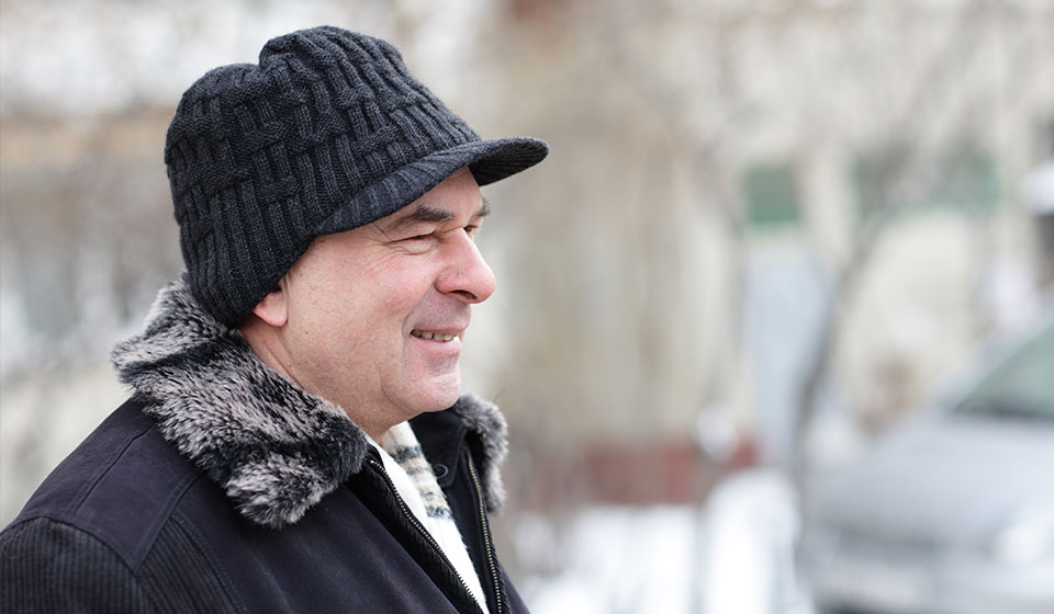 Homme portant un manteau avec col en fourrure et un bonnet noir, debout à l’extérieur par temps froid.