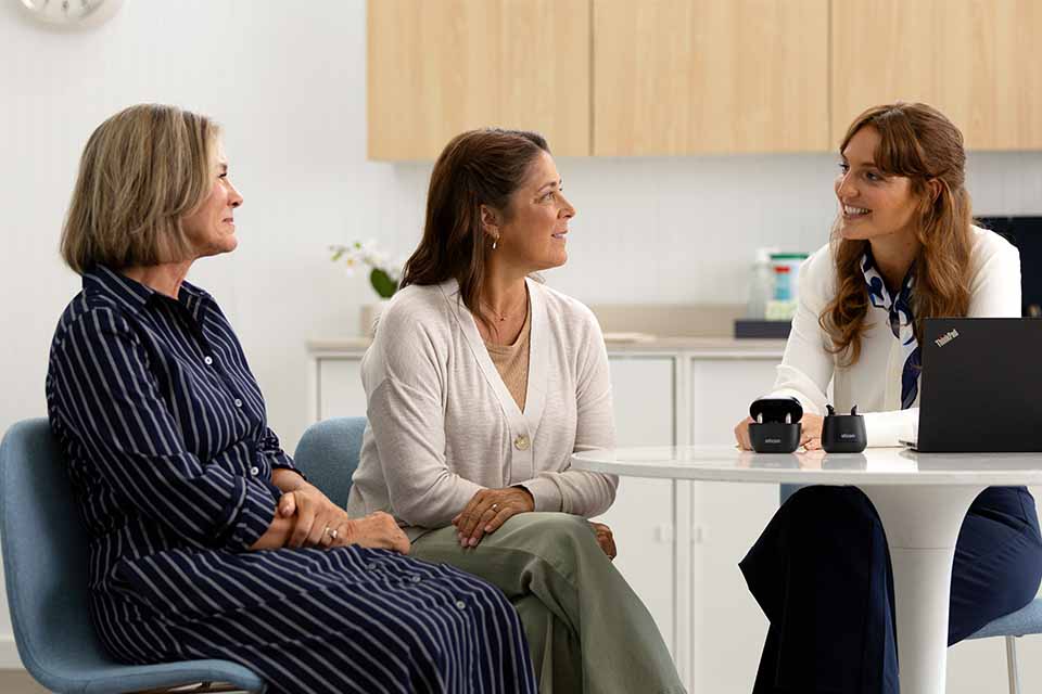 Deux femmes consultent une audiologiste dans une clinique Audika lumineuse. Sur la table, un ordinateur portable Lenovo se trouve près de boîtiers Audika contenant probablement des appareils auditifs.