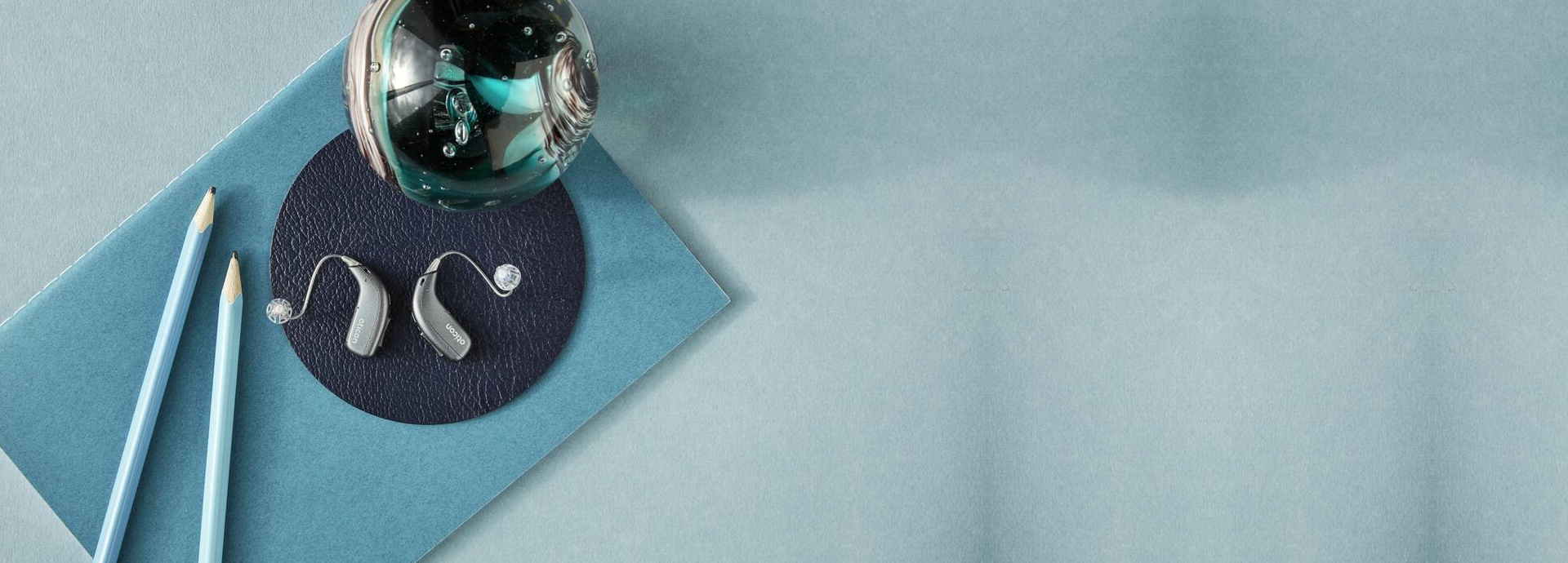 Deux appareils auditifs modernes Oticon posés sur une surface circulaire en cuir, accompagnés de crayons bleus et d’un presse-papier décoratif, sur un fond bleu élégant.
