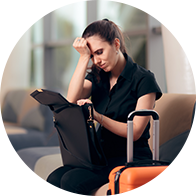 Une femme assise dans un salon tenant sa tête d’une main et fouillant dans son sac avec une valise posée à côté d’elle.