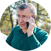 Un homme avec des cheveux gris utilisant un téléphone à l'extérieur, entouré d'arbres.