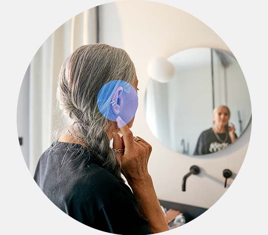 Une femme aux cheveux gris ajuste son appareil auditif devant un miroir dans une salle de bain moderne éclairée, reflétant une atmosphère calme et intime. Aucun texte visible.