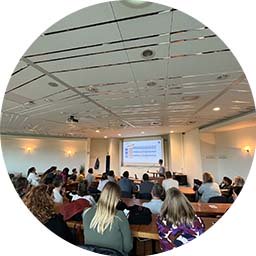 Un auditoire assis regarde une présentation projetée dans une salle de conférence moderne, avec des plafonniers lumineux et un conférencier debout à l’avant. Aucun texte identifiable.