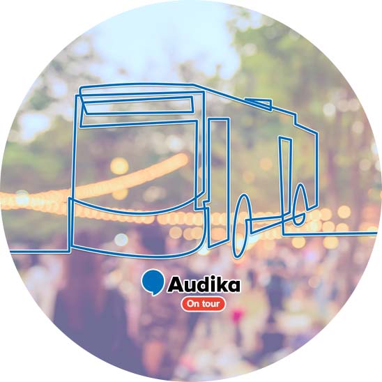 Un bus stylisé en ligne bleue est superposé sur un arrière-plan flou d’un événement en extérieur avec guirlandes lumineuses. Texte : « Audika - On tour ».