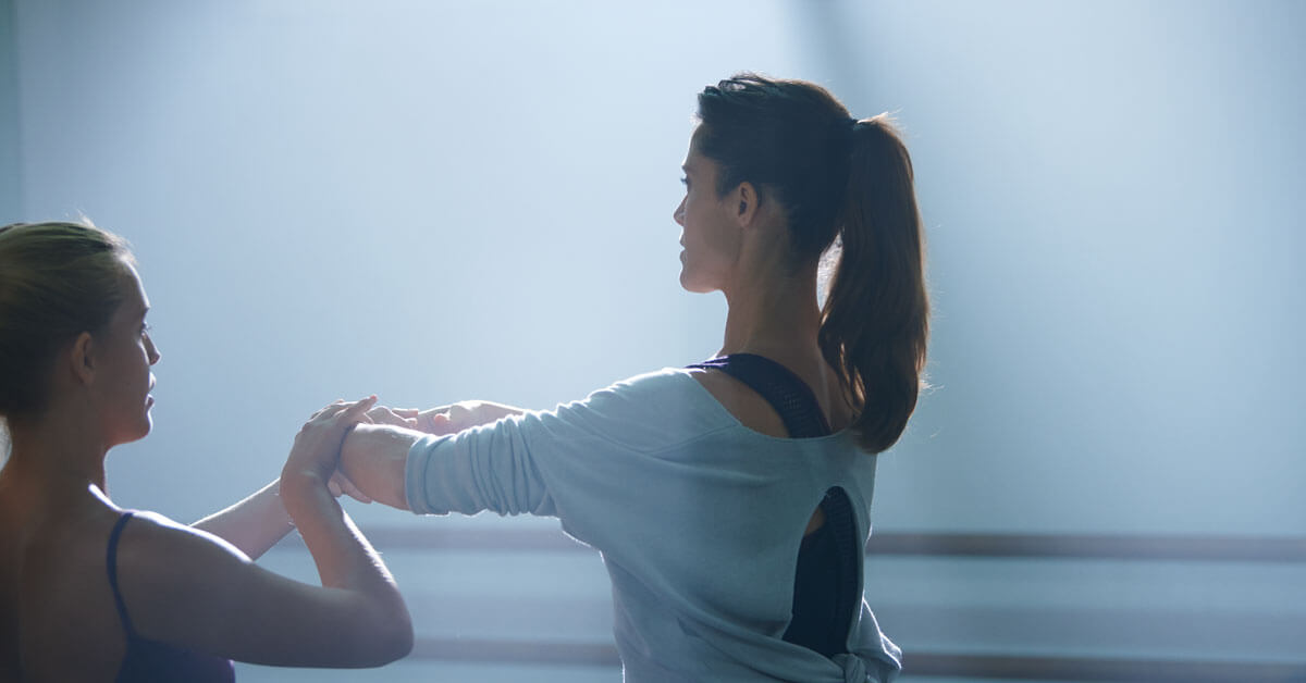 Zwei Frauen in einem Tanzstudio üben eine Tanzbewegung. Eine Frau trägt ein hellblaues Oberteil mit offenem Rücken. Sonnenlicht fällt durch Fenster und beleuchtet den Raum. Kein Text sichtbar.